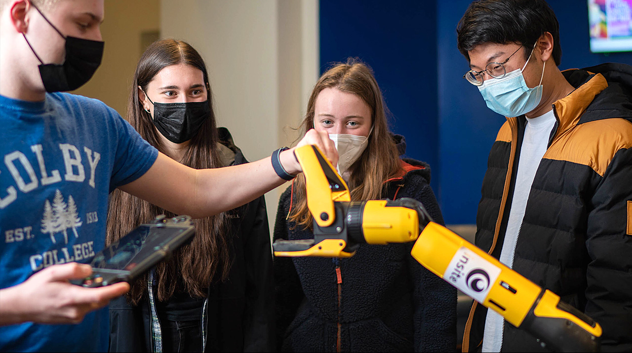 Students observe a robotic arm being demonstrated, wearing masks in an indoor setting.