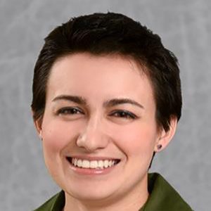 Professional headshot of a woman with short dark hair, smiling against a gray background, wearing a green shirt and blazer.