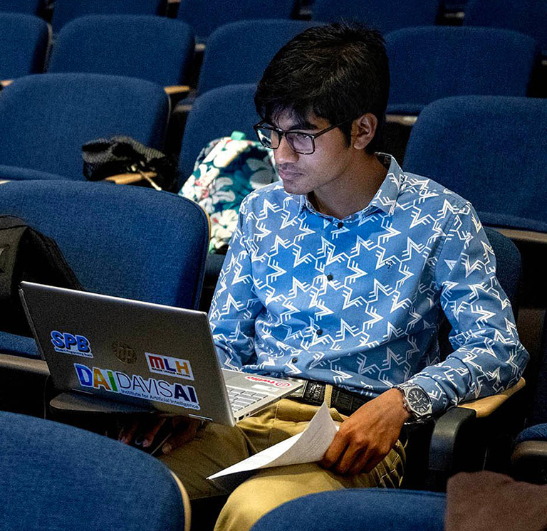 A student working on a laptop in an auditorium, wearing a patterned shirt and surrounded by empty chairs.
