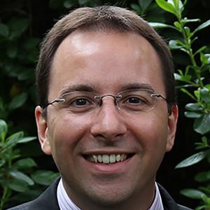 A smiling man in a suit with glasses, standing outdoors among greenery.
