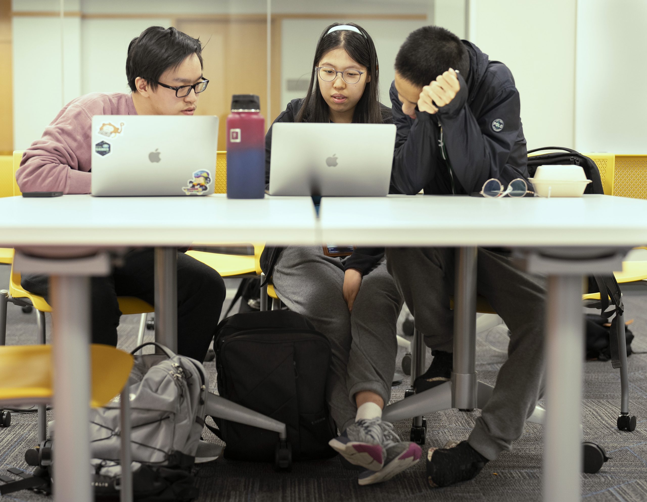 Three students working together at a table, using laptops and studying in a bright, modern classroom setting.