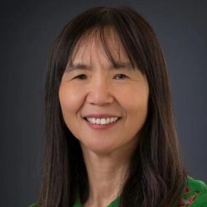 Portrait of a woman with long dark hair, smiling, wearing a green top with colorful embroidery on a dark background.