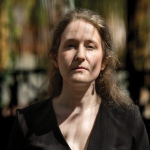 Woman with long hair wearing a black top, gazing directly at the camera with a blurred background.