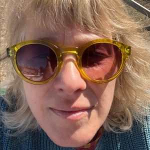 Person with blonde hair wearing yellow sunglasses, smiling, against an outdoor background. Bright and sunny day.