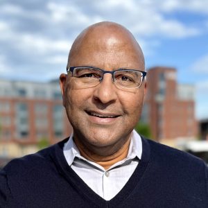 Smiling man with glasses, wearing a dark sweater over a collared shirt, posing outdoors with buildings in the background.