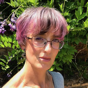 Woman with short, pink-highlighted hair, wearing glasses, smiling among green foliage and purple flowers.