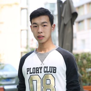 A young man wearing a PLORY CLUB 08 sweatshirt stands outdoors, surrounded by greenery and buildings in the background.