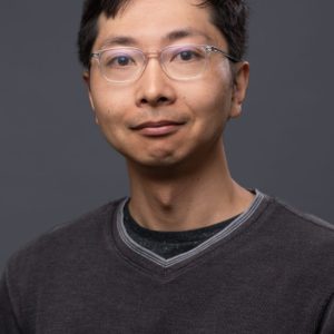 Portrait of a man with glasses wearing a dark sweater, looking directly at the camera against a gray background.