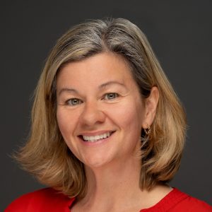 Smiling woman with shoulder-length blonde hair and a red top, against a dark background.