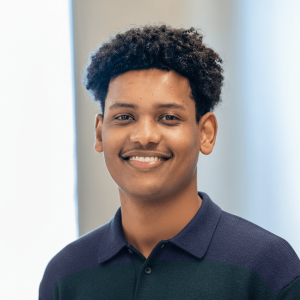 Smiling young man with curly hair, wearing a dark polo shirt, against a light background, conveying a friendly demeanor.