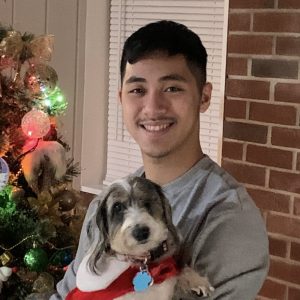 Young man smiling while holding a small dog, next to a decorated Christmas tree with colorful lights.