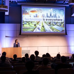 Person speaking on stage with a large screen showing a cityscape and the text Technology is everywhere, in an audience setting.