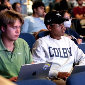 Two students focused on laptops during a lecture, seated in an auditorium with an engaged audience in the background.