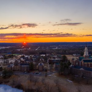 Aerial view of a campus at sunset, featuring buildings and a lake, with vibrant orange and purple sky.